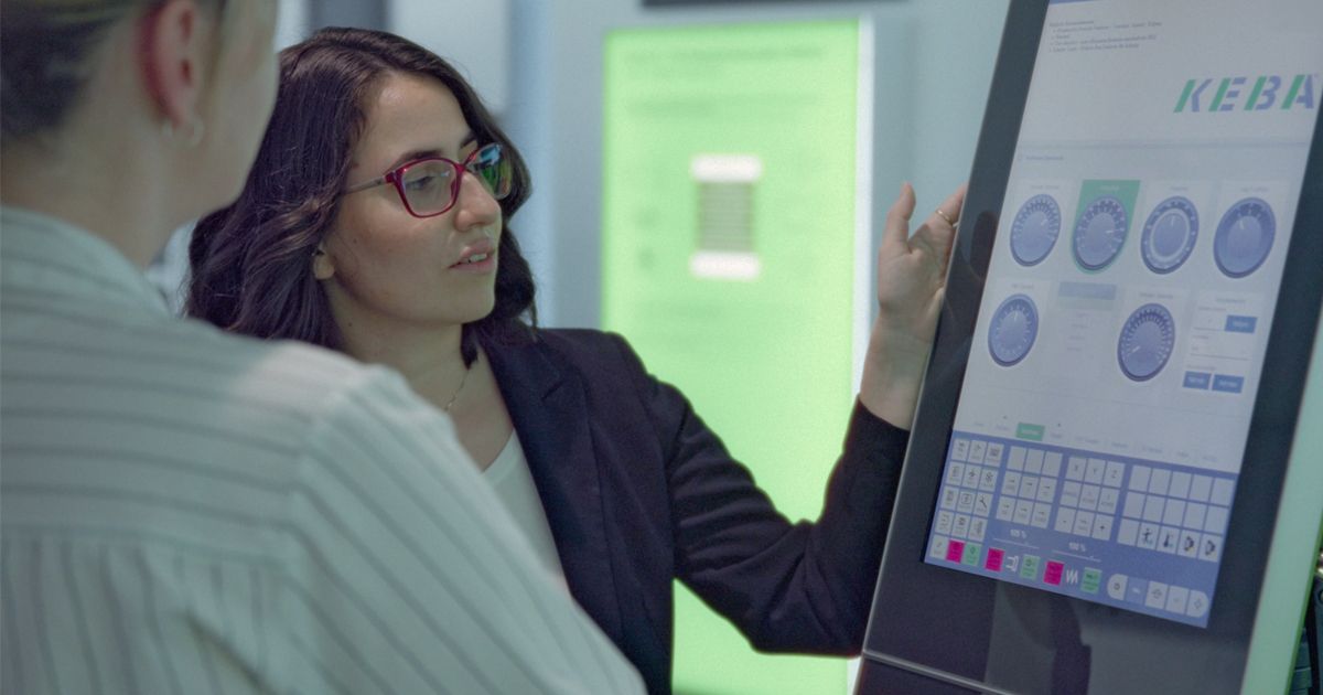 Eine Mitarbeiterin erklärt einer Kollegin eine industrielle Steuerung über ein modernes Touchpanel mit der Benutzeroberfläche von KEBA.