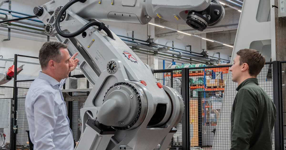 autonox operations manager Clemens Ratajczak (left) and a colleague (right) stand in front of a large industrial robot arm in a factory hall; one person points to the robot part, the other listens.