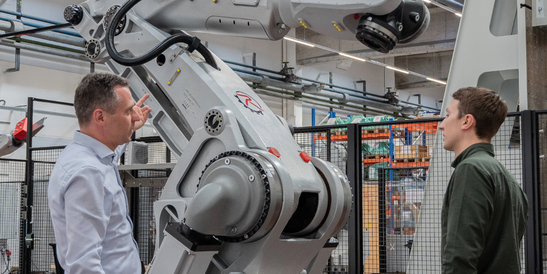 autonox operations manager Clemens Ratajczak (left) and a colleague (right) stand in front of a large industrial robot arm in a factory hall; one person points to the robot part, the other listens.
