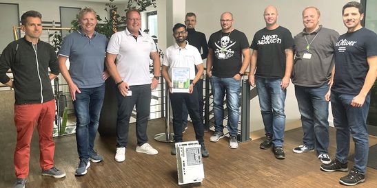 Eine Gruppe von neun KEBA-Teammitgliedern aus Unna und Indien steht im Foyer zusammen, während die Person in der Mitte eine Auszeichnung für eine erfolgreich absolvierte Schulung präsentiert. Vor ihnen befindet sich ein PitchOne-Gerät, das den Bezug zur Automatisierungstechnologie und der internationalen Zusammenarbeit symbolisiert.