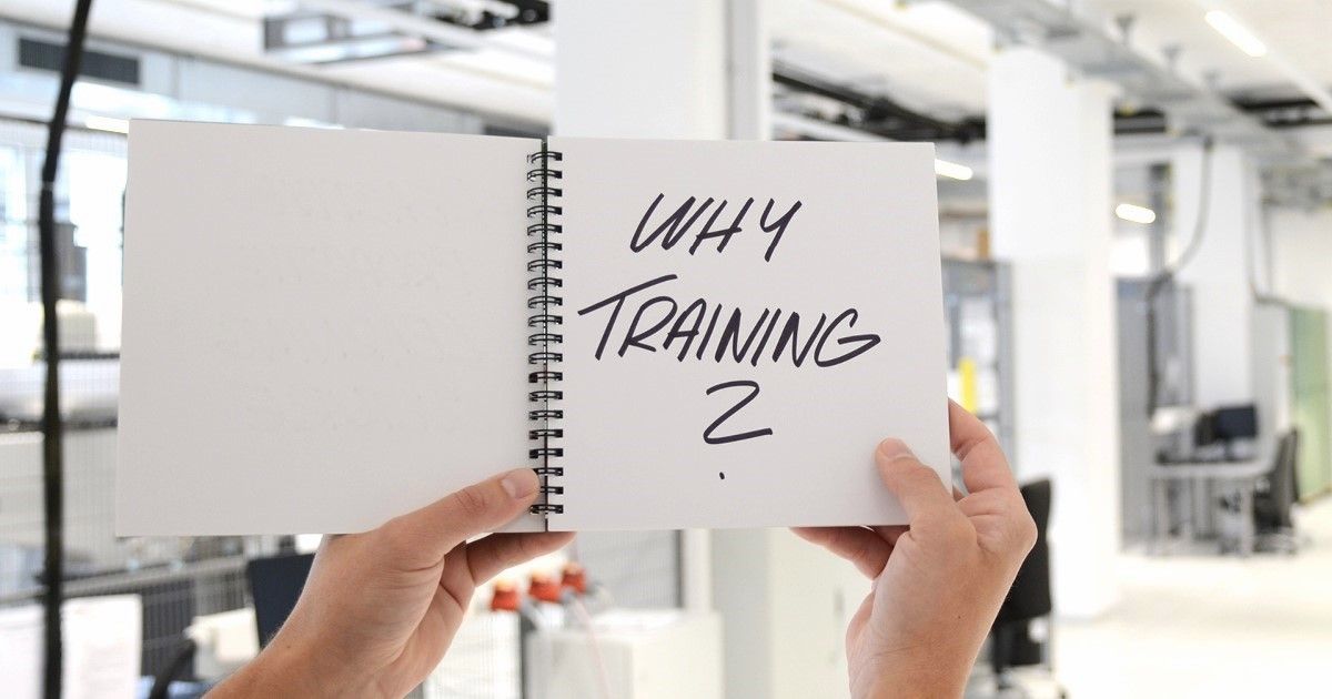 An engineer holding an open spiral notebook in an office or workshop environment. On the right page, handwritten text reads “WHY TRAINING?” while the left page is blank.