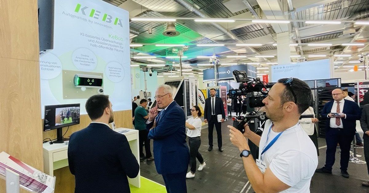 Visitors at the 2025 SicherheitsExpo interacting at the KEBA exhibition booth, discussing the AI-based monitoring system KeBob, while a camera crew films the presentation.