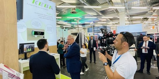 Besucher auf der SicherheitsExpo 2025 am KEBA-Messestand im Gespräch über das KI-gestützte Überwachungssystem KeBob, während ein Kamerateam die Präsentation filmt.