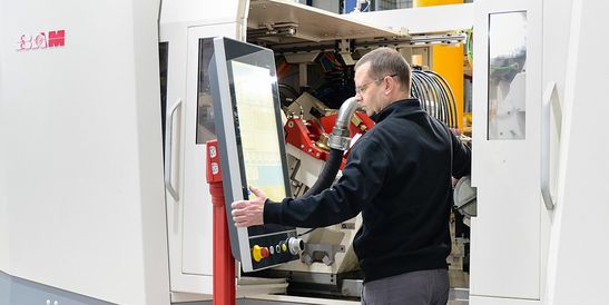 An operator stands in front of a large machine and uses a swivel-mounted HMI panel for control. The system is designed as an industrial machining center.