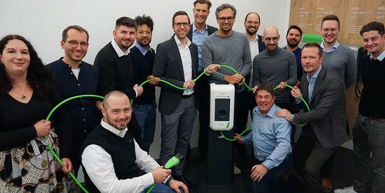 A group of KEBA and The Mobility House employees gathered around a wallbox, holding a green charging cable – symbolizing the ten-year partnership between the two companies.