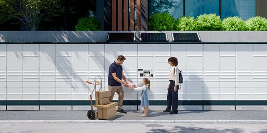 A family receives a package from a delivery person in front of a modern building with lockers and greenery.