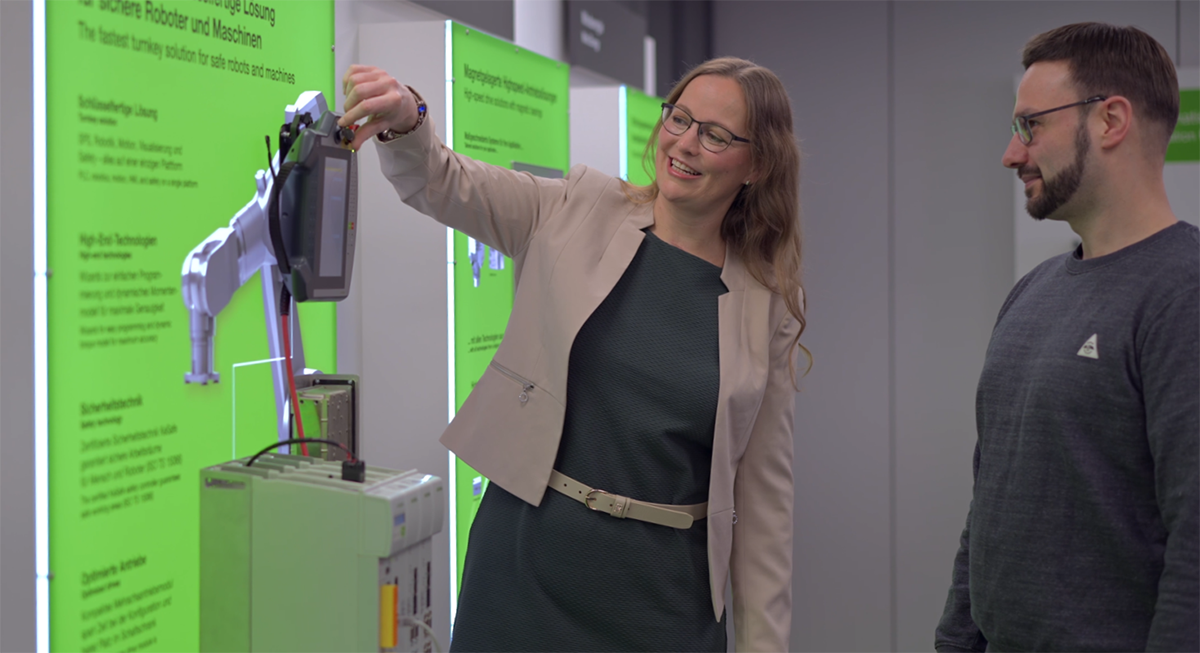 Eine Mitarbeiterin demonstriert einem Kollegen ein Handbediengerät an einem Robotik-Exponat. Im Hintergrund befinden sich grüne Informationstafeln zu Robotern und Maschinen.