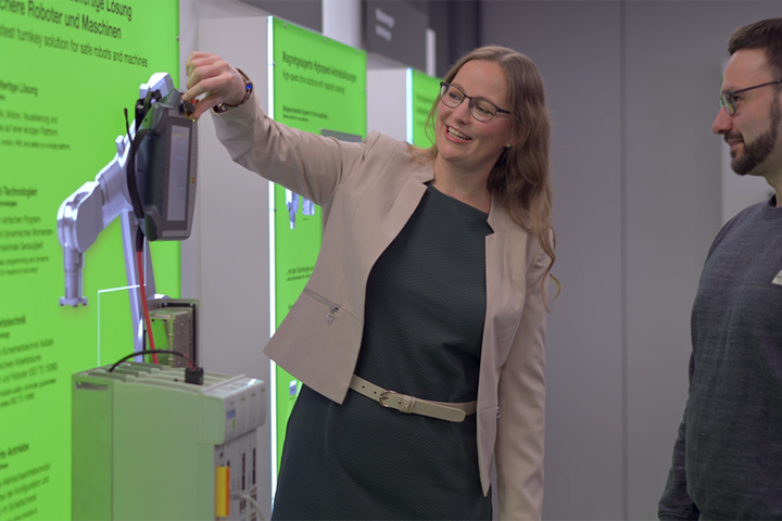 Eine Mitarbeiterin demonstriert einem Kollegen ein Handbediengerät an einem Robotik-Exponat. Im Hintergrund befinden sich grüne Informationstafeln zu Robotern und Maschinen.