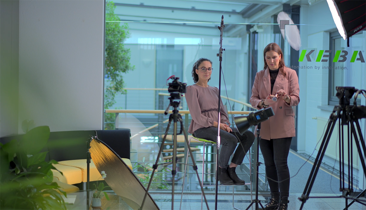 Zwei Mitarbeiterinnen befinden sich in einem für Foto- oder Videoaufnahmen eingerichteten Bereich. Eine sitzt vor der Kamera, während die andere neben ihr die Ausrüstung vorbereitet.