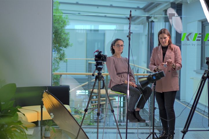 Zwei Mitarbeiterinnen befinden sich in einem für Foto- oder Videoaufnahmen eingerichteten Bereich. Eine sitzt vor der Kamera, während die andere neben ihr die Ausrüstung vorbereitet.