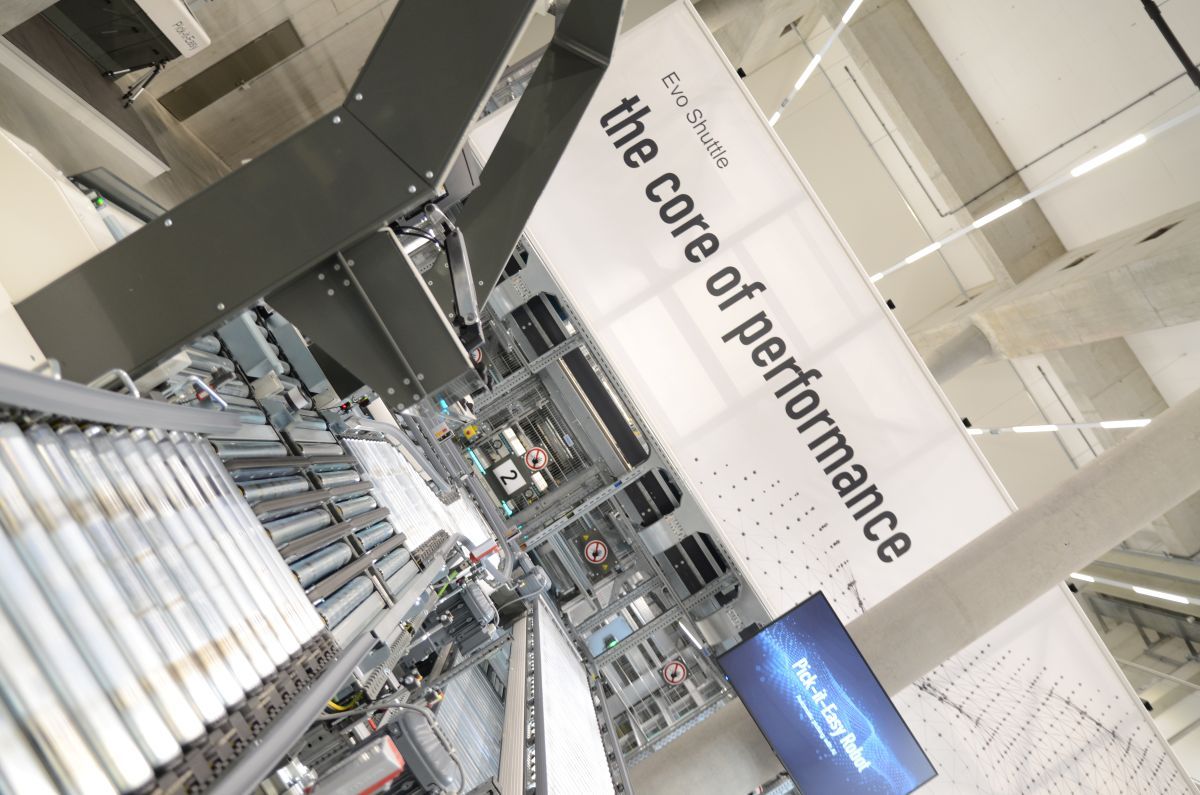 Industrial machinery with conveyor belts in a modern facility. A sign reads "Evo Shuttle: the core of performance" in the background.