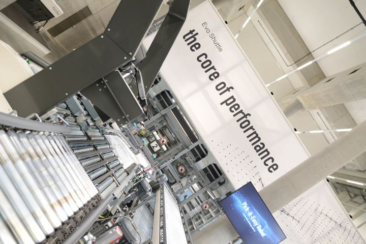 Industrial machinery with conveyor belts in a modern facility. A sign reads "Evo Shuttle: the core of performance" in the background.