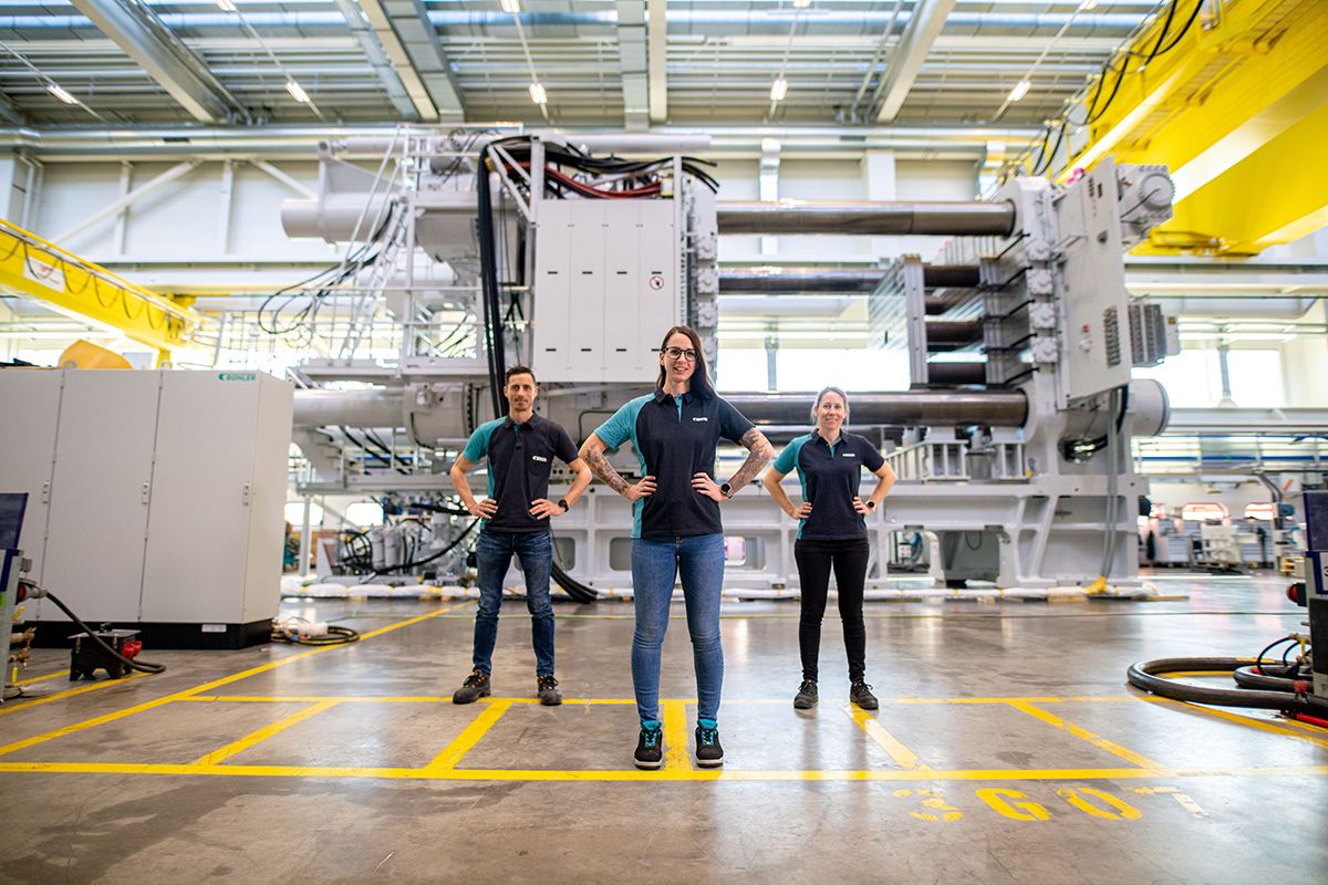 Drei Buehler-Mitarbeiter:innen in Arbeitsuniformen stehen selbstbewusst in einer großen Industrieanlage mit Maschinen im Hintergrund.