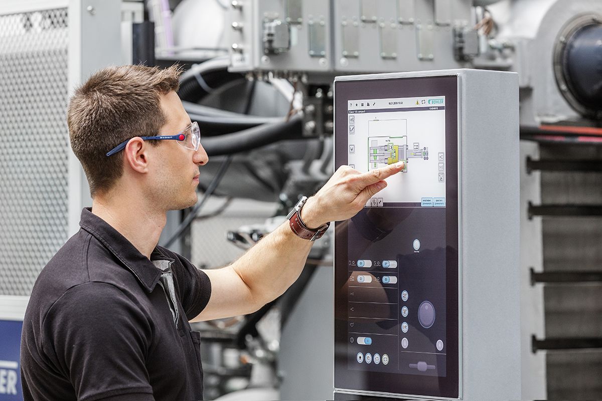 Ein Mann mit Schutzbrille und schwarzem Hemd, der eine Touchscreen-Schnittstelle an einer Industriemaschine bedient, in einer modernen Fabrikumgebung.