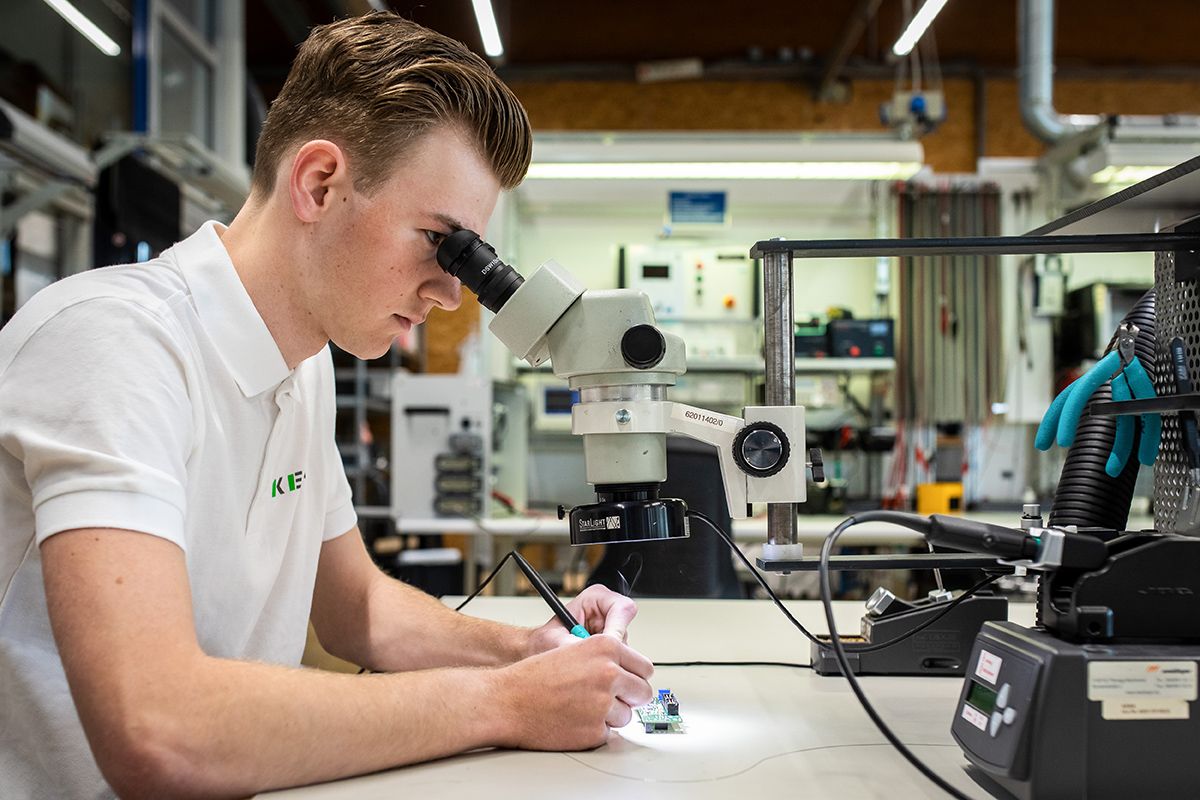 Ein KEBA-Auszubildender inspiziert eine Leiterplatte unter dem Mikroskop und führt präzise Lötarbeiten in einem professionellen Elektroniklabor durch.