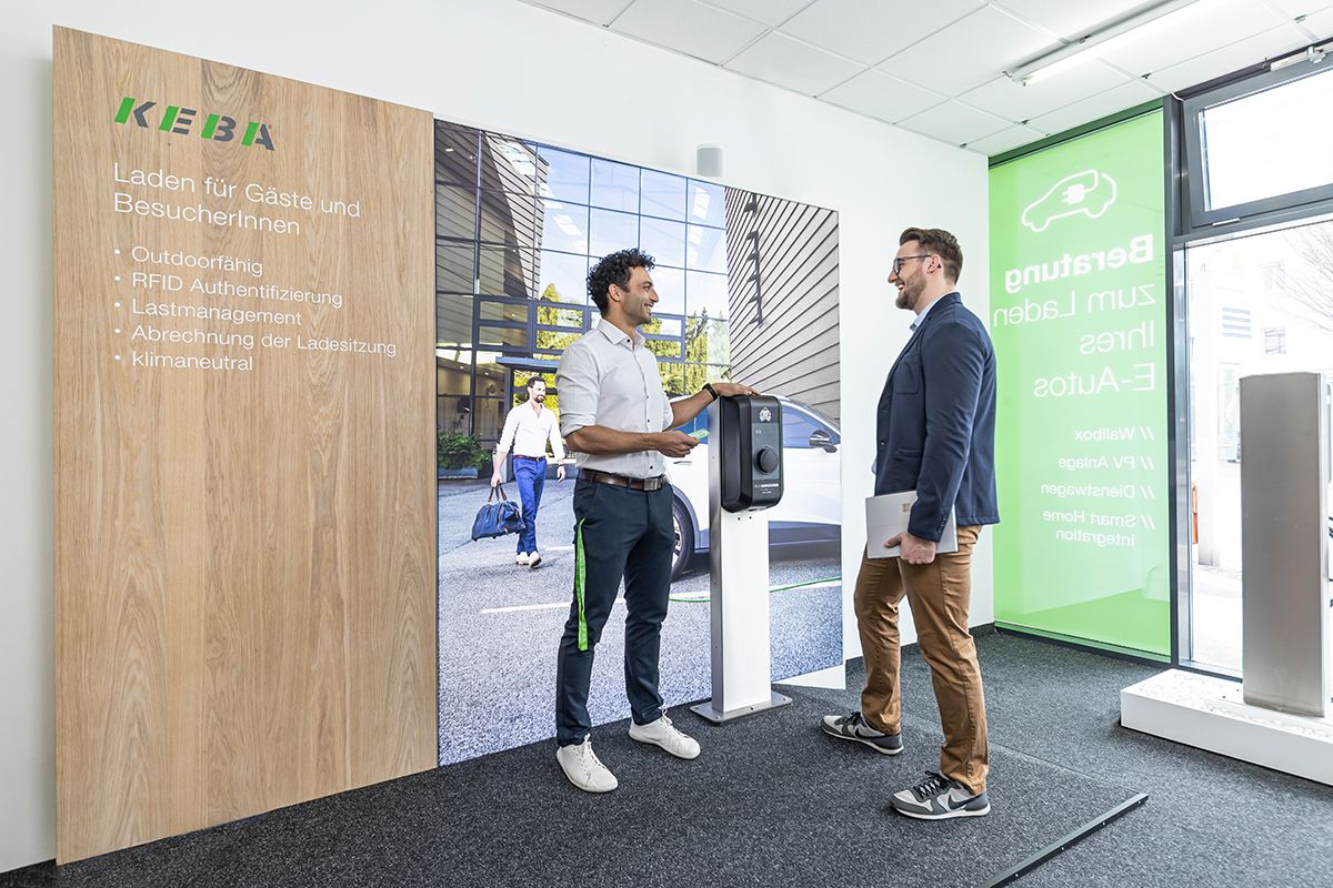 Two men in conversation in front of a KEBA wallbox and info panel for guests and visitors.
