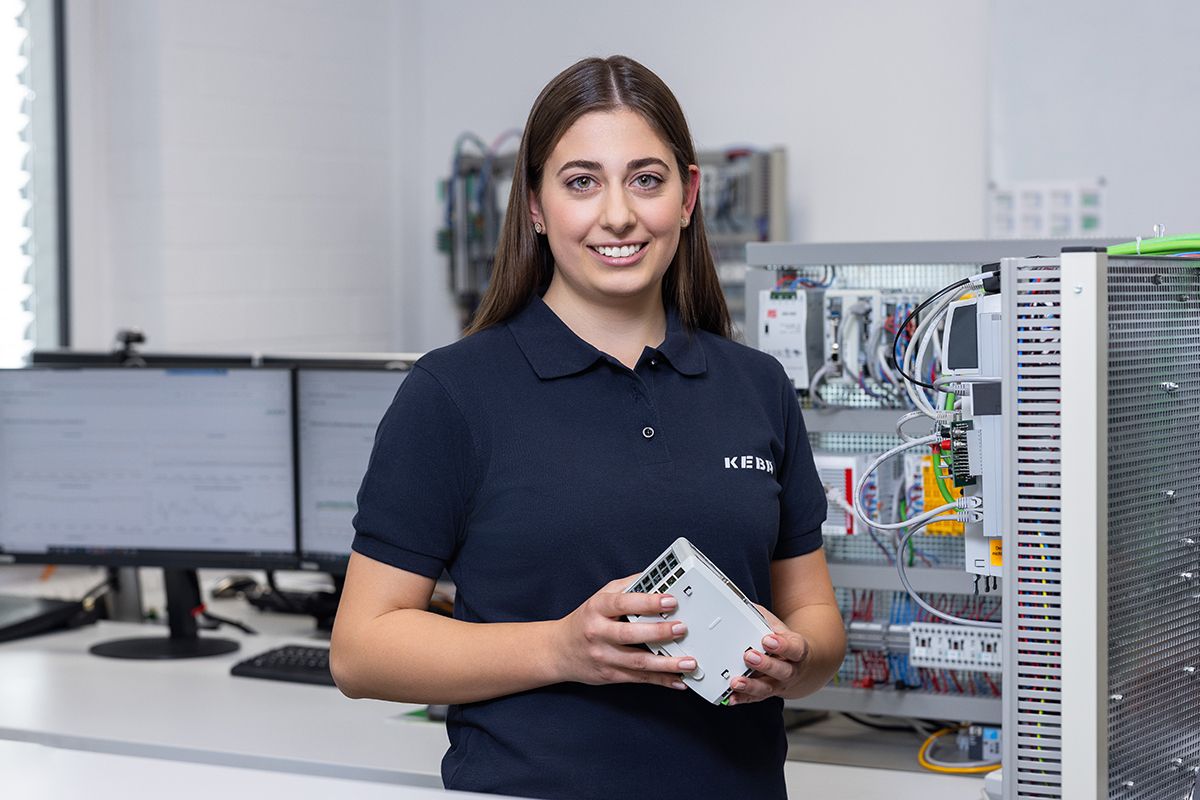 Eine KEBA-Mitarbeiterin präsentiert ein Steuerungsmodul vor einem Schaltschrank – Technikkompetenz im Fokus.