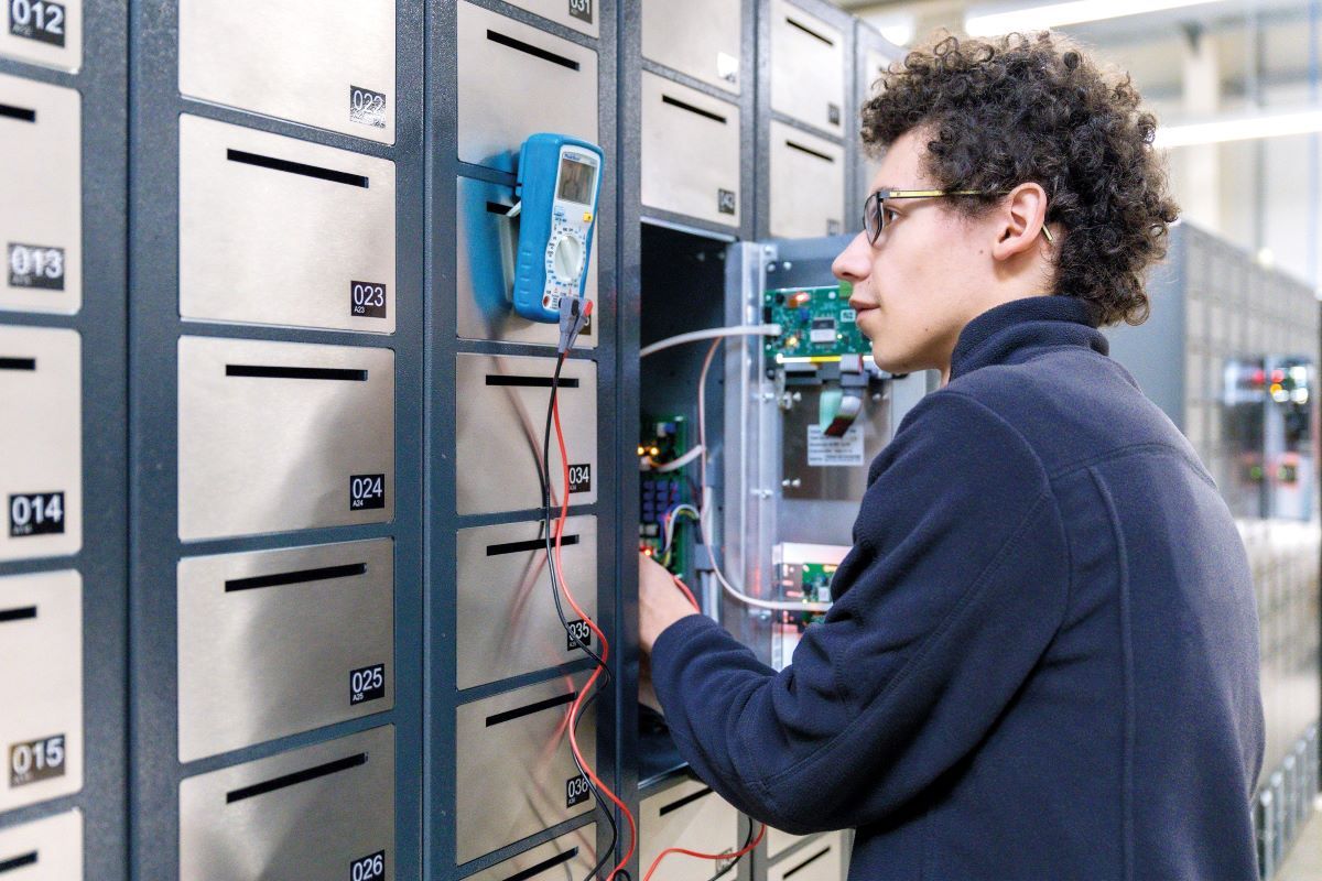 Ein KEBA-Mitarbeiter überprüft mit einem Messgerät die Elektronik eines geöffneten Schließfachsystems in einer Reihe von Metallfächern.