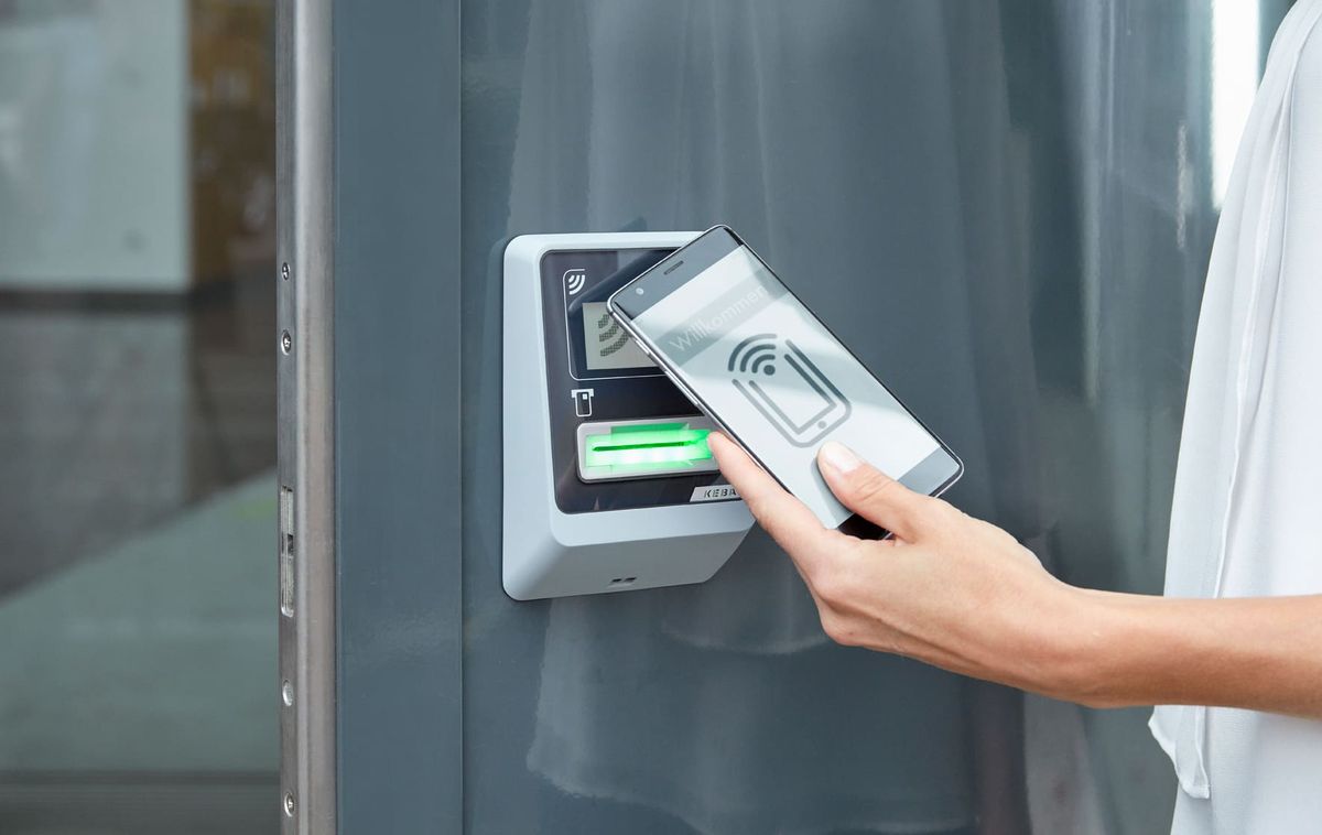 Person nutzt ein Smartphone, um ein sicheres Zugangssystem mit kontaktlosem Kartenleser an der Wand zu bedienen.