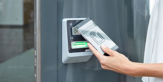 Person using a smartphone to access a secure entry system with a contactless card reader on a wall.