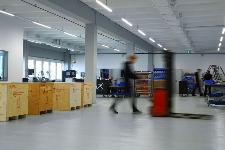 Interior view of a bright industrial hall: On the left are several labeled wooden crates, in the middle a person is moving a load with a pallet truck, and in the background two people are working at workstations with tool trolleys and machines.