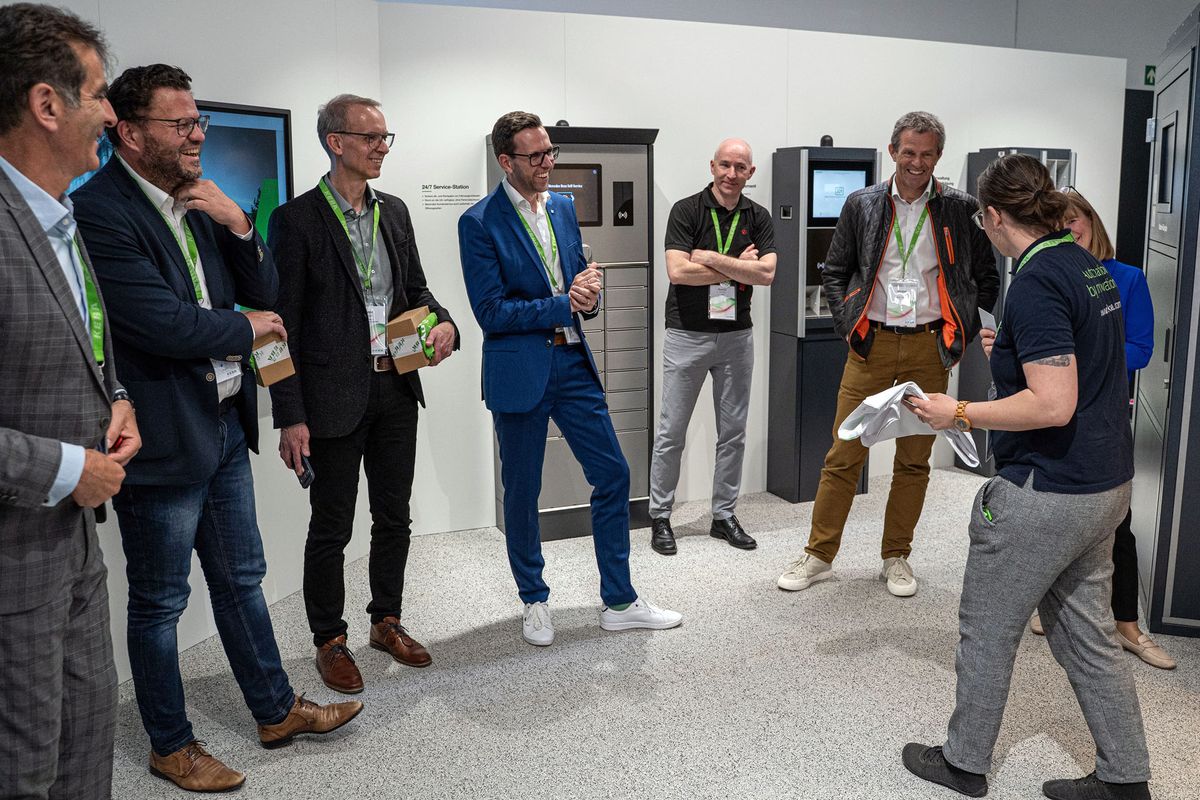 A group of people stand in a KeMas showroom, some laughing and casually dressed. A person in front holds a white shirt, engaging the group's attention.