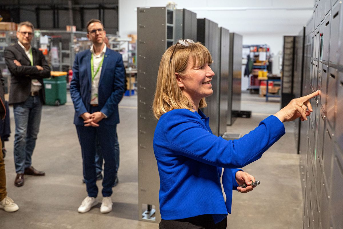 A woman in a blue jacket points at a KeMas locker with three observing men in the background.