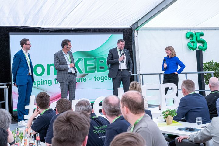 Four people stand on stage speaking at an event, with an audience seated in front and a banner displaying "35" in the background.