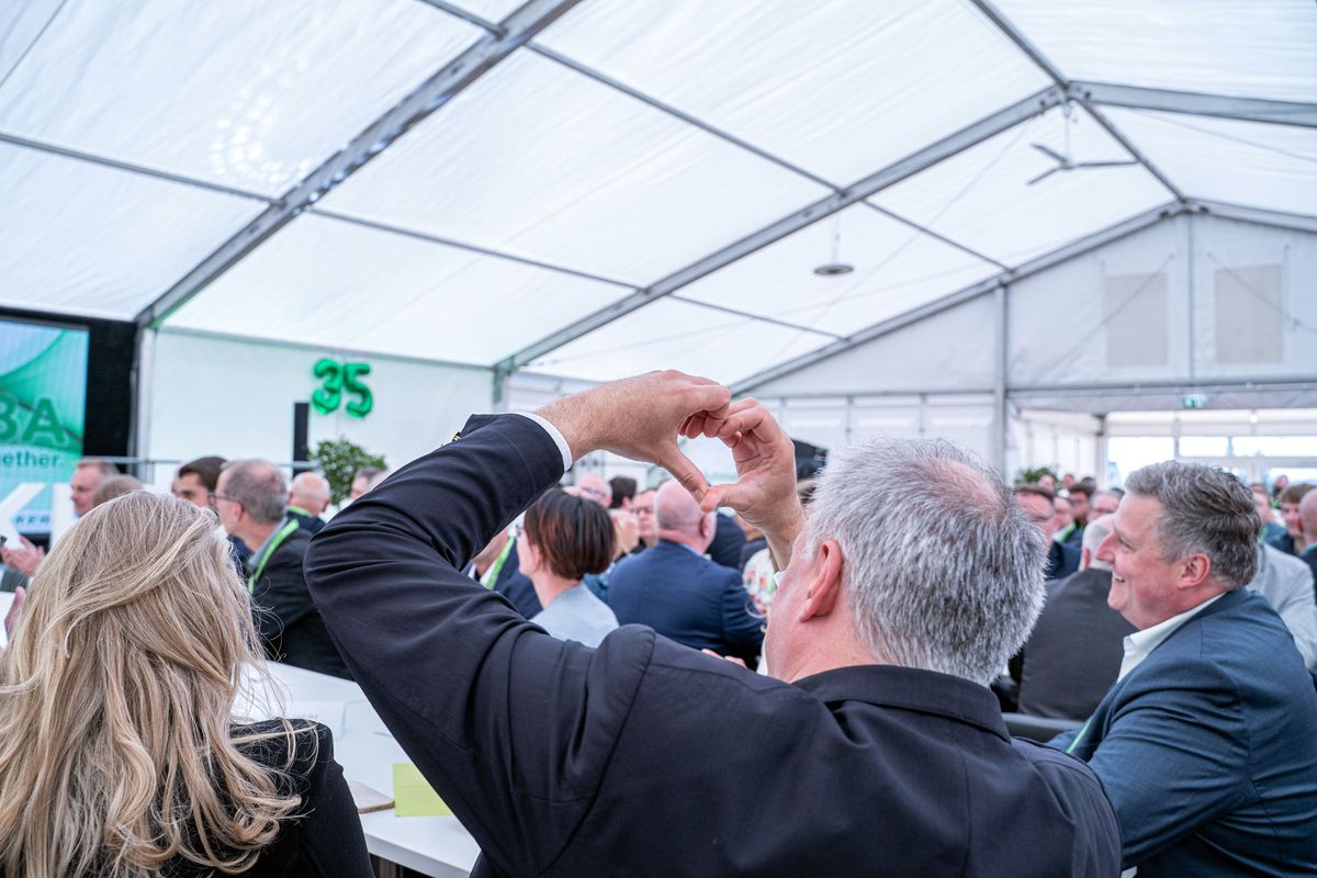 A person forms a heart shape with their hands at a crowded business event inside a large white tent.