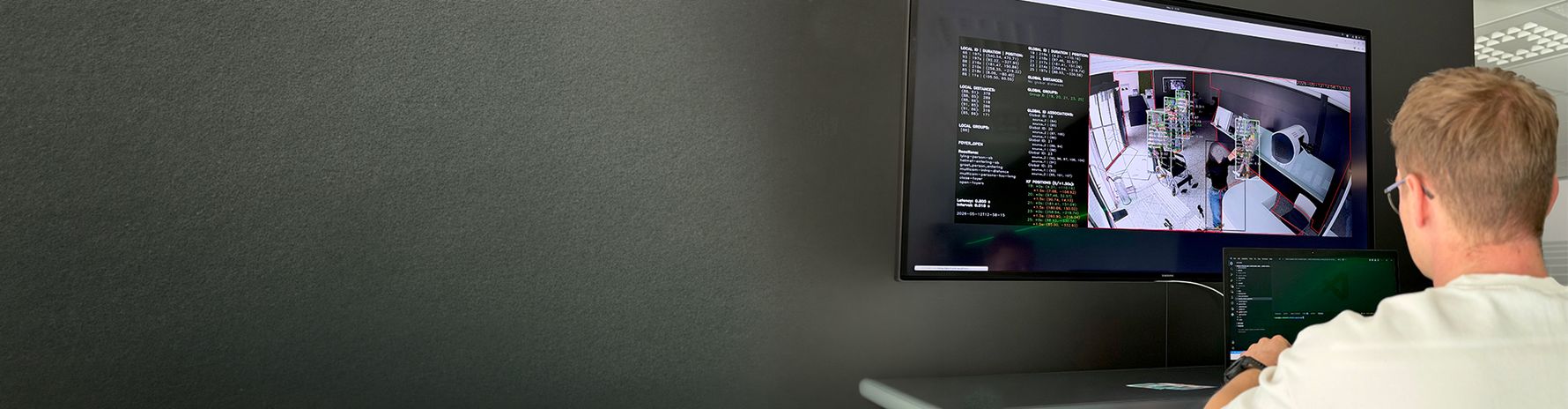 Ein Mitarbeiter sitzt vor einem Laptop und einem großen Monitor. Auf dem Monitor ist eine Kameraaufnahme eines Raums mit visuellen Markierungen und technischen Daten zur Erkennung und Positionsbestimmung von Personen und Objekten zu sehen.
