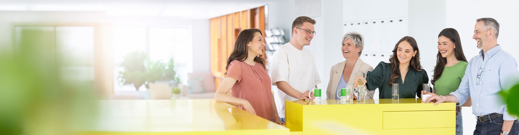Six colleagues stand together at a yellow kitchen counter in a bright office, talking over drinks.