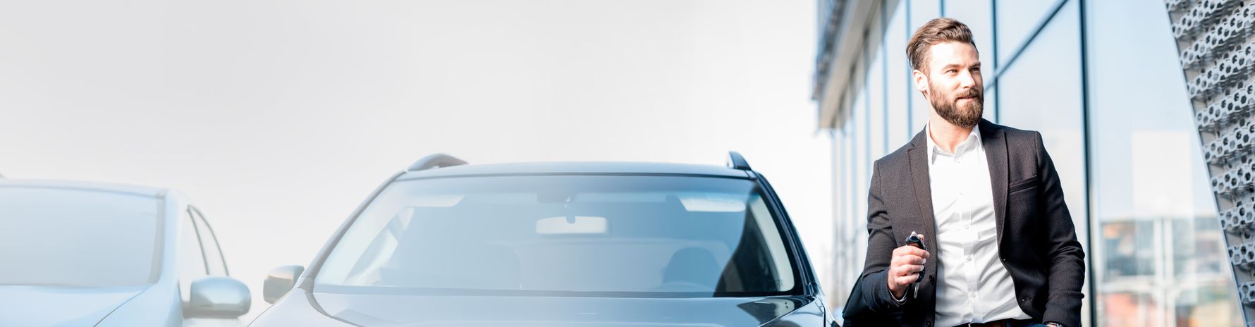 A man in a suit is standing next to a car, holding car keys in his hand, with a modern building in the background.