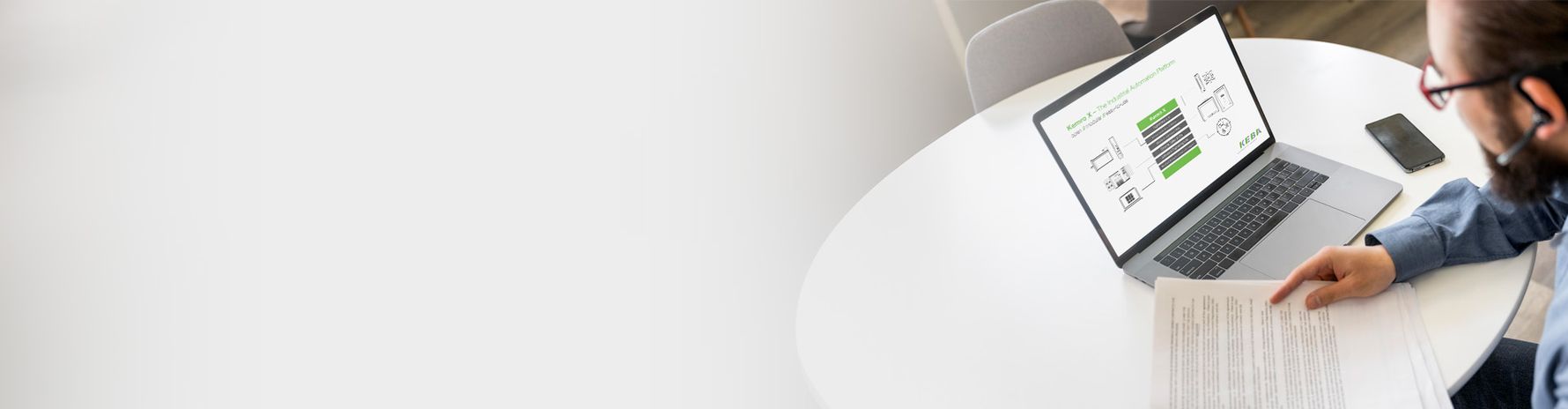 Eine Person sitzt an einem runden Tisch und arbeitet mit einem Laptop, auf dessen Bildschirm eine schematische Automatisierungs- oder Systemübersicht mit KEBA-Bezug angezeigt ist. In der Hand hält die Person ein Dokument, daneben liegt ein Smartphone. Das Bild steht für digitale Beratung, Projektabstimmung und Remote-Services in der industriellen Automatisierung.