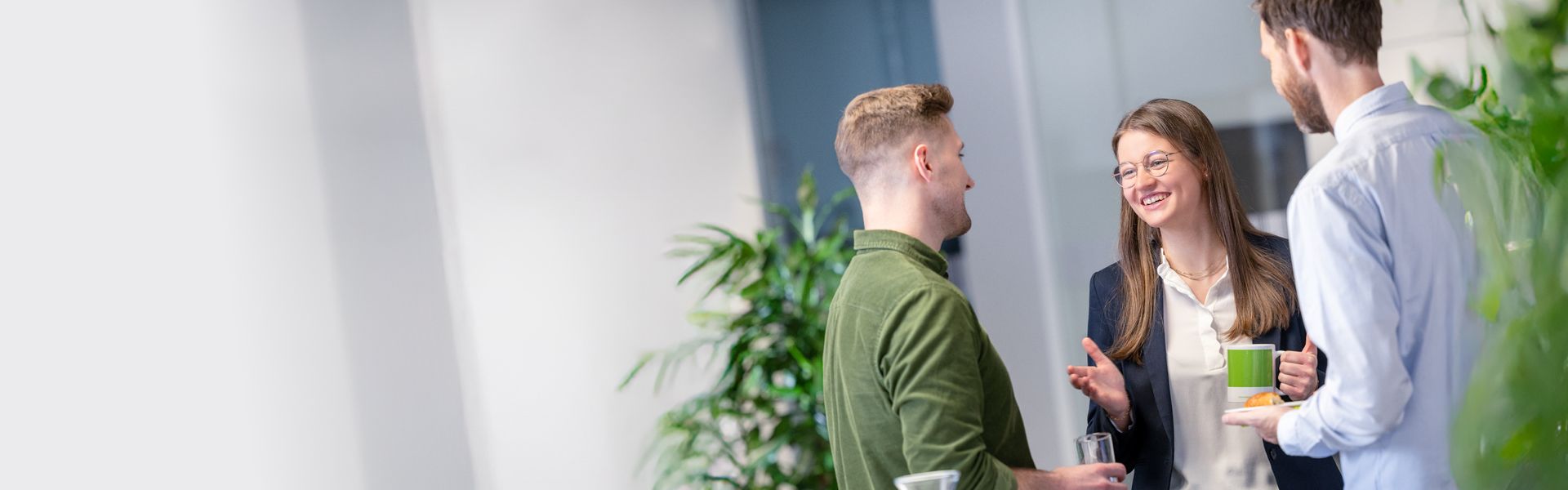 Three colleagues are standing together in a modern office environment, engaged in a friendly conversation. Plants and bright lighting in the background create a fresh and welcoming workspace.