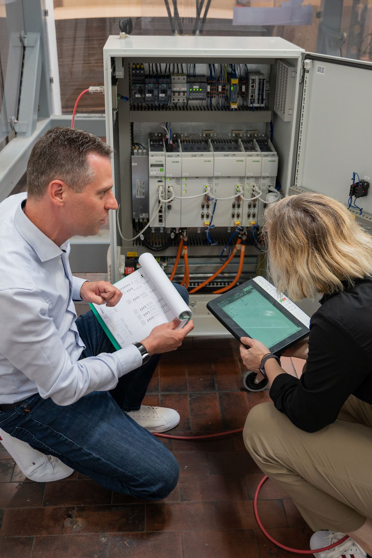 Betriebsleiter Clemens Ratajczak und eine Kollegin prüfen einen geöffneten KEBA-Schaltschrank: Eine Person hält Schaltpläne in der Hand, die andere bedient ein KeTop.