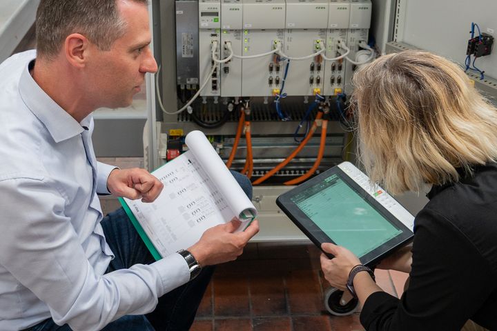 Operations manager Clemens Ratajczak and a colleague inspect an open KEBA control cabinet: one person holds circuit diagrams in their hand, while the other operates a KeTop.