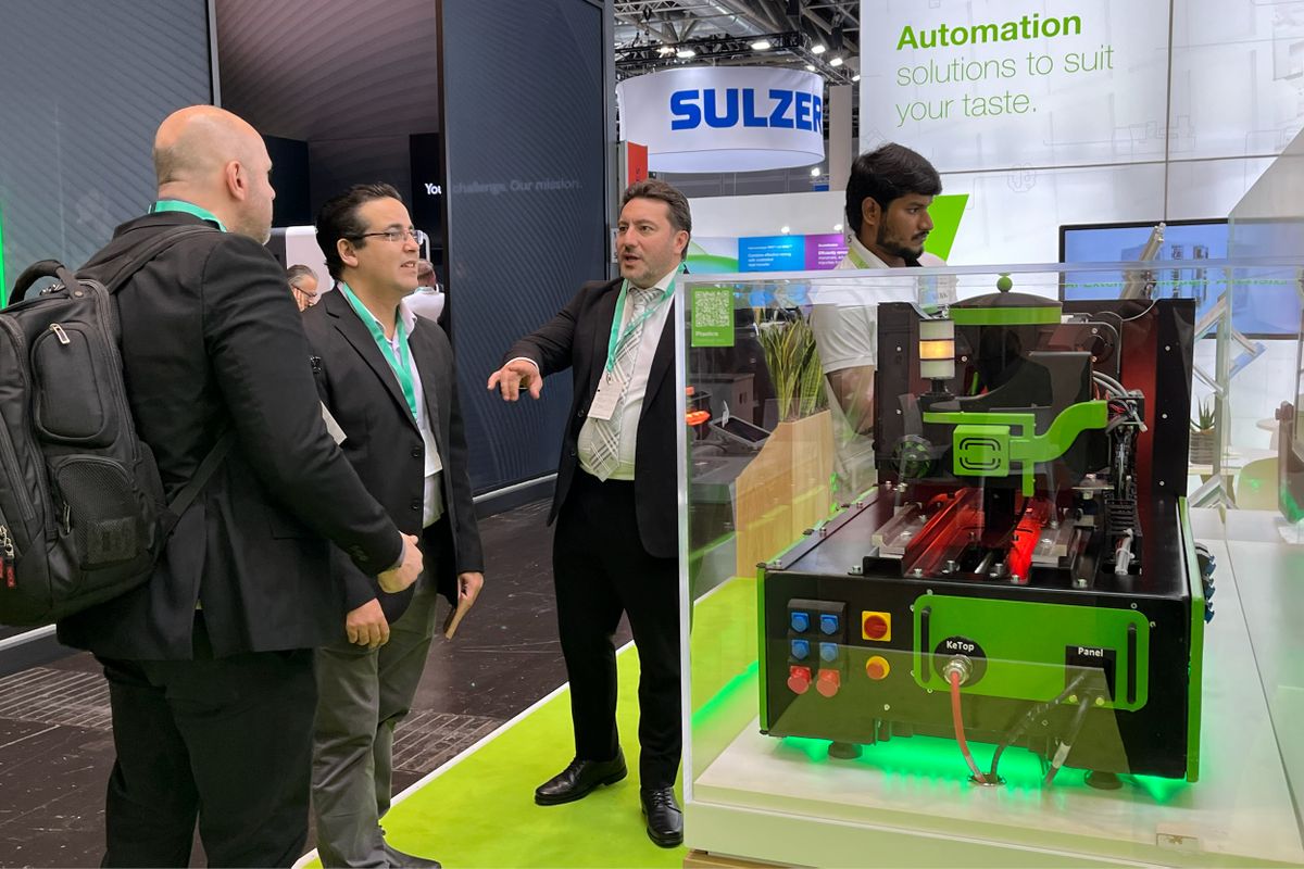 KEBA representative explaining a machine demo of KePlast IMM Simulatro to visitors at the booth. The KEBA branding and slogan “Automation solutions to suit your taste” are visible in the background.