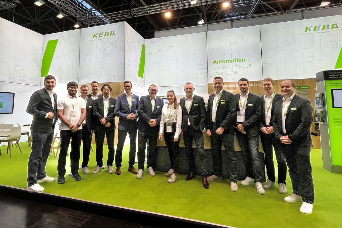 Group photo of KEBA team members standing together at a trade fair booth. The background shows the KEBA exhibition stand with white and green branding and the slogan “Automation solutions to suit your taste.” All team members are wearing business attire with KEBA badges, smiling at the camera.