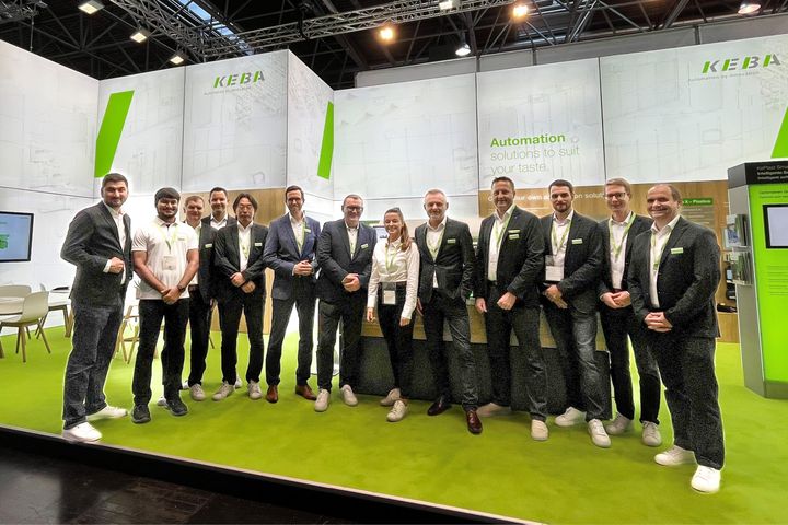 Group photo of KEBA team members standing together at a trade fair booth. The background shows the KEBA exhibition stand with white and green branding and the slogan “Automation solutions to suit your taste.” All team members are wearing business attire with KEBA badges, smiling at the camera.