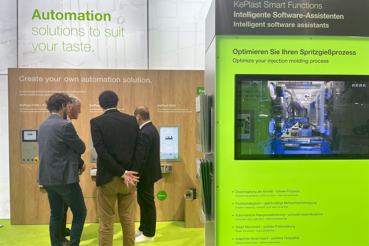 Several trade fair visitors engaged in discussion at KEBA’s automation display area. A green-lit exhibit labeled “Optimize your injection molding process” showcases KEBA’s KePlast Smart Functions for intelligent software-assisted production.