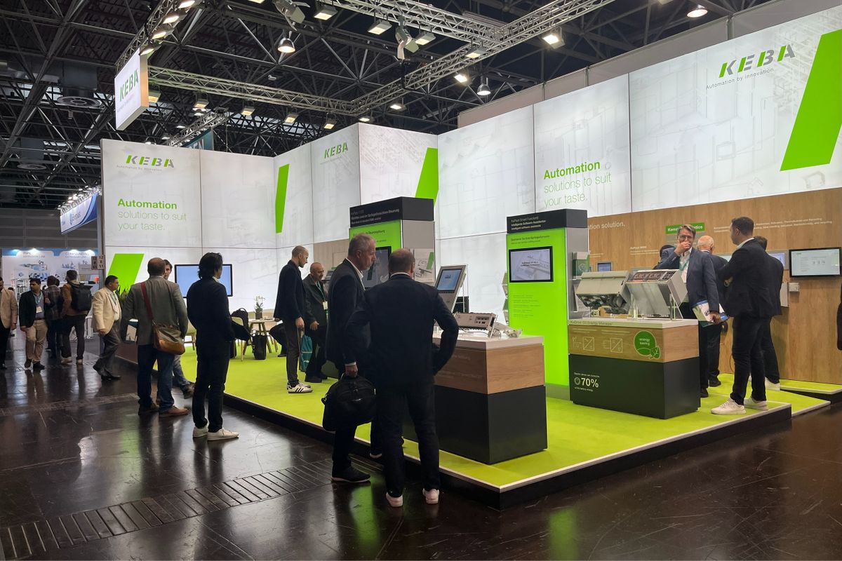 Visitors and KEBA representatives interacting at the KEBA trade fair booth. The booth features bright green flooring, modern displays, and product demos. The KEBA logo and slogan “Automation solutions to suit your taste” are prominently visible on the backdrop.