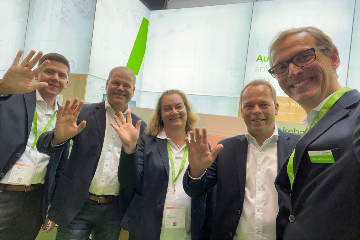 Five KEBA team members posing and waving at the camera in front of the company’s trade fair booth. All are dressed in business attire with green KEBA lanyards, smiling warmly. The booth design in the background features KEBA’s signature green and white branding.