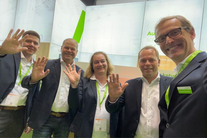 Five KEBA team members posing and waving at the camera in front of the company’s trade fair booth. All are dressed in business attire with green KEBA lanyards, smiling warmly. The booth design in the background features KEBA’s signature green and white branding.