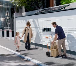 Stație modernă de colete KEBA KePol Loxmate în utilizare – o femeie cu un copil și un curier interacționează cu echipamentul.