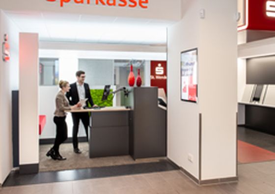 Due persone interagiscono presso un moderno sportello bancario con l’insegna “Sparkasse”, con schermi digitali e ATM visibili in un ambiente luminoso e minimalista.