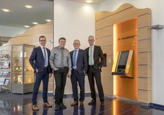 Quattro uomini in abito da lavoro sorridono nell’atrio di un ufficio, accanto a un cash recycler di KEBA.