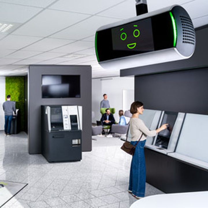 Eine moderne Bankfiliale mit Menschen, die Bankautomaten bedienen werden vom KI-Assistent KeBob bewacht.