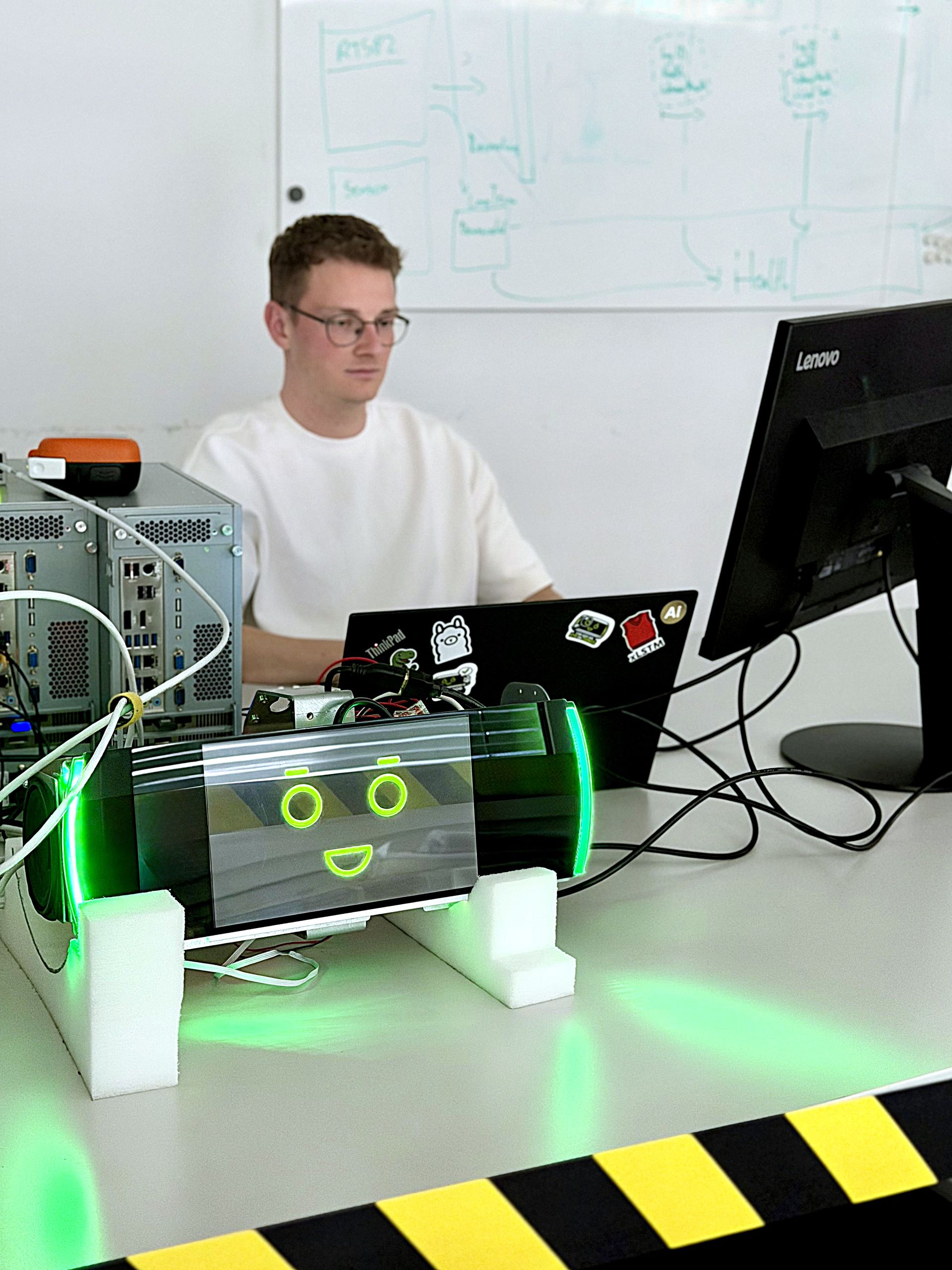 Person with glasses works at a computer workstation with several devices and cables. In the foreground is a technical device with a green illuminated smiling face.