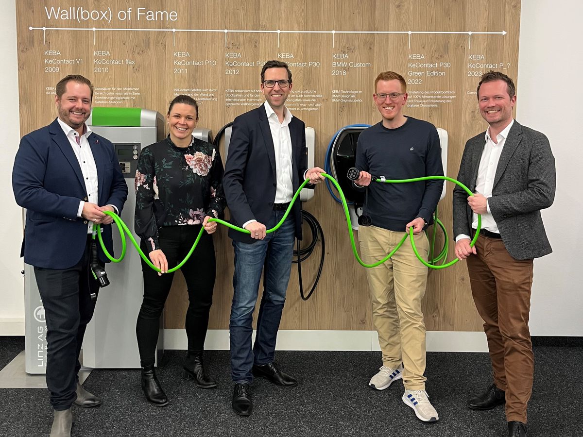 Four people holding a green charging cable together in front of a wall displaying various KEBA wallbox models.