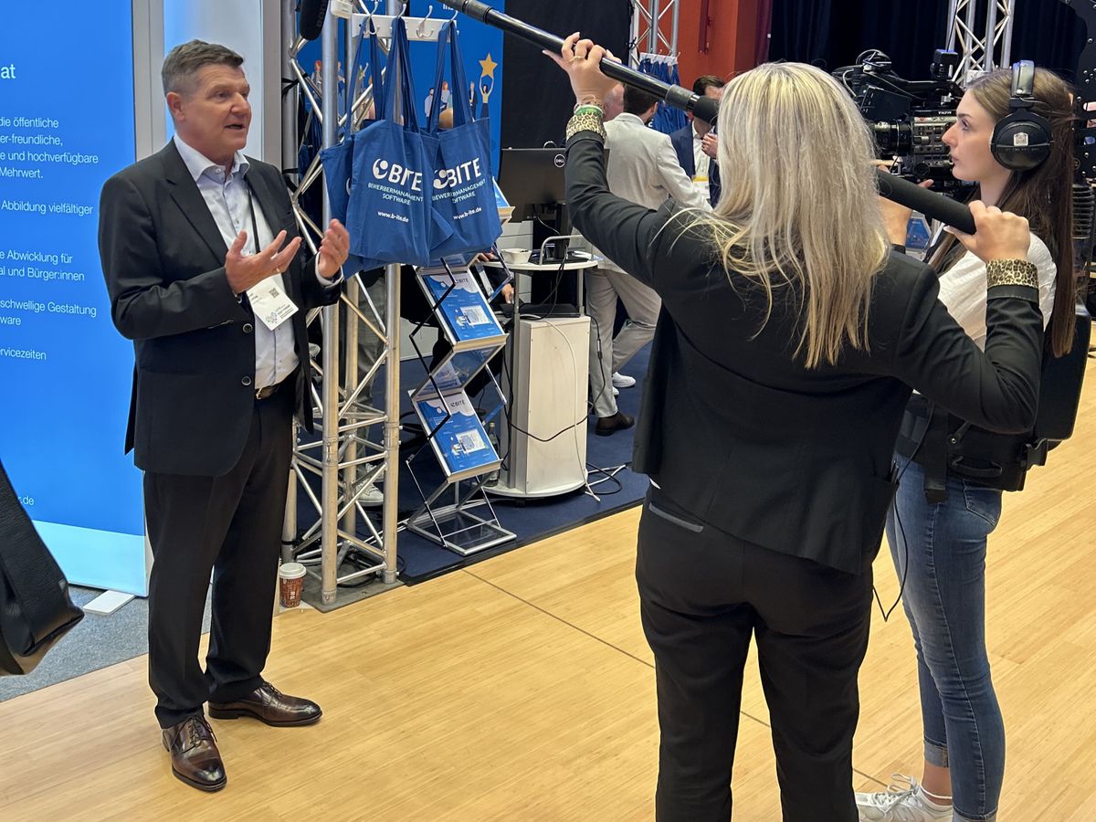 Ein Mann am Messestand wird von zwei Reporterinnen interviewed.